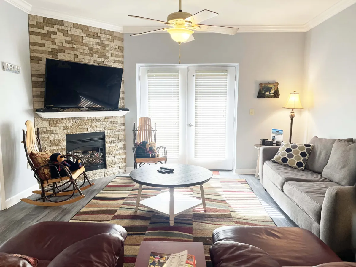 This image captures a spacious living area with a stone fireplace, flat-screen TV, round coffee table, and sliding glass doors that frame an inspiring view. The comfortable seating arrangement invites relaxation and socializing, and the mountain scenery is subtly visible through the doors.