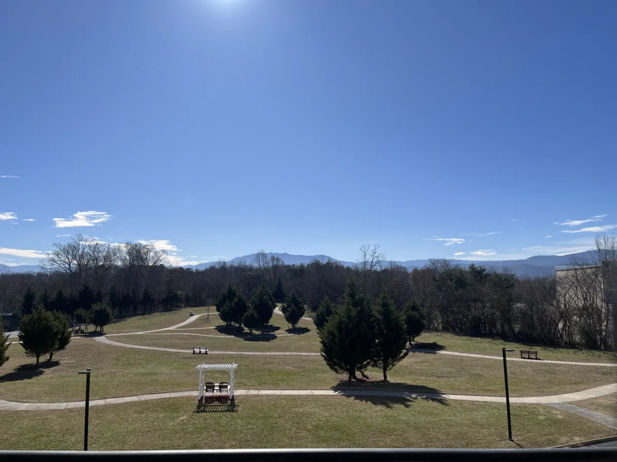 A scenic walking trail with green grass, pathways, benches, and a charming gazebo, framed by mountains in the distance. The area is inviting for walks, gatherings, and outdoor leisure.