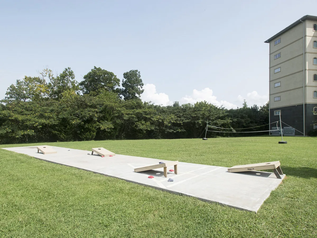 A grassy outdoor space featuring cornhole boards set up for play, with green lawns and a building in the background. The scene is casual and relaxed, suggesting opportunities for friendly games, picnics, and outdoor socializing.