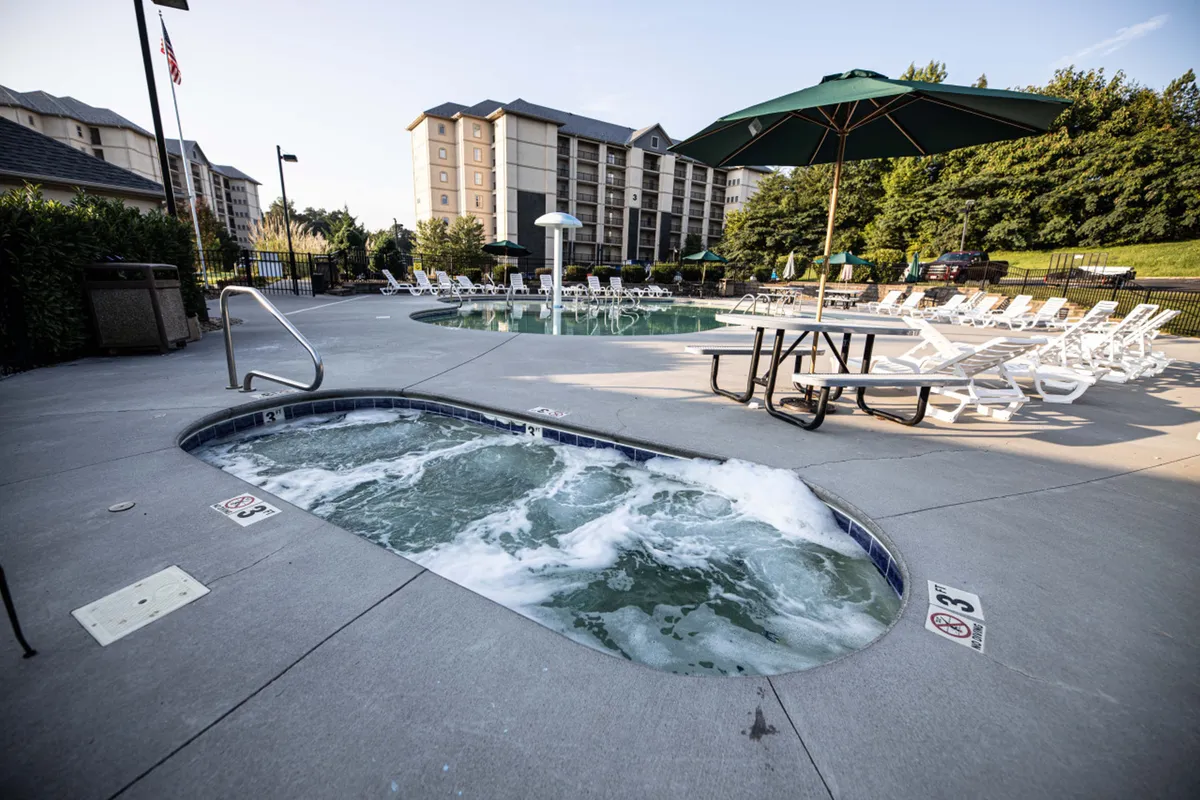 A spacious outdoor pool area featuring a glistening pool, a hot tub, multiple lounge chairs, and an umbrella-shaded table. The setting is sunny and inviting, ideal for both energetic swimming and relaxed socializing by the water.