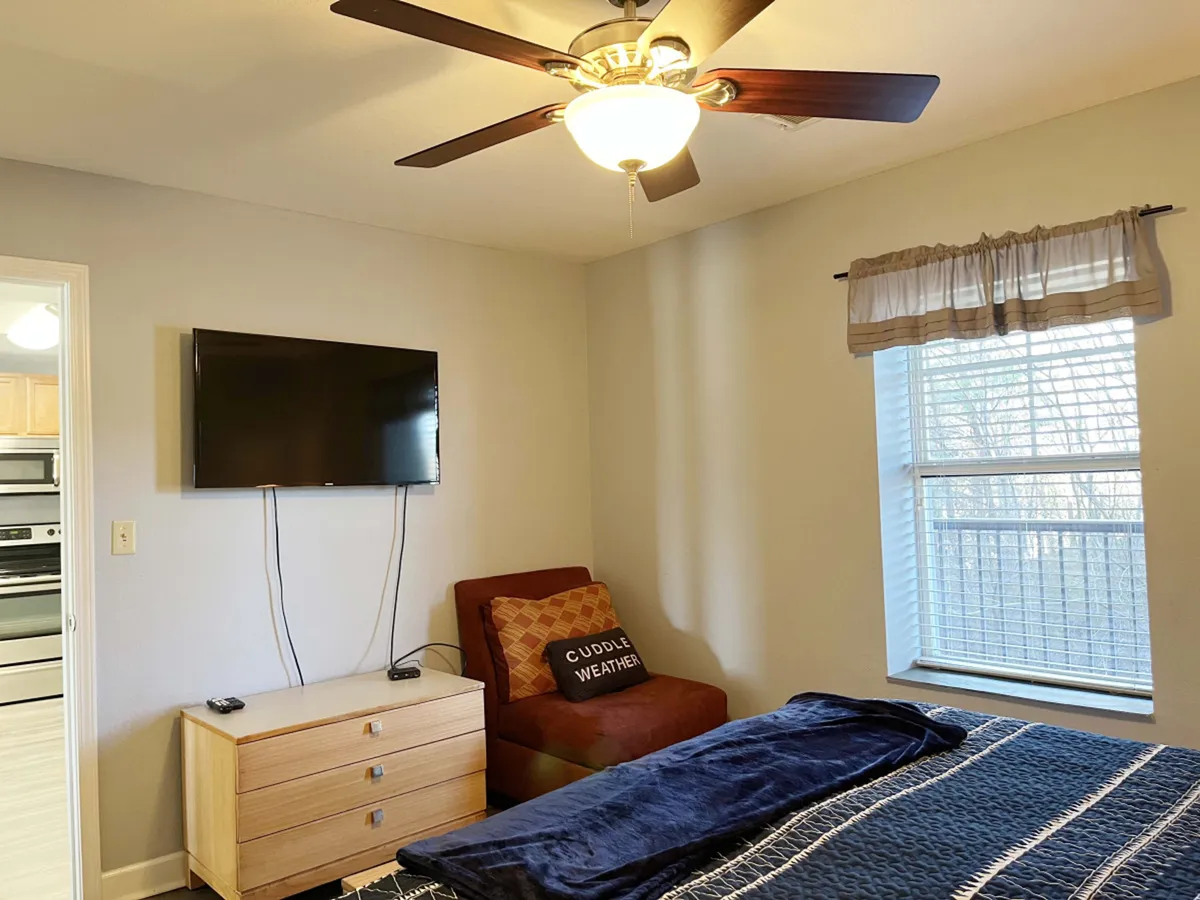 An inviting bedroom with a king-sized bed dressed in a blue cover, a cheerful red chair sporting a 'Cuddle Weather' pillow, and a wall-mounted television. The setup creates a perfect space for relaxing with a favorite show or unwinding after a day of adventure.