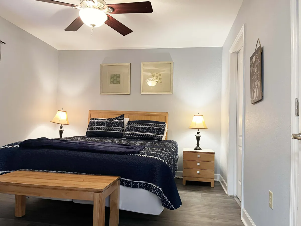 A serene and cozy bedroom featuring a king-sized bed adorned with plush bedding, flanked by nightstands and soft-glowing lamps. Artwork on the wall adds a personal touch, while the room's tranquil ambiance invites relaxation and restful sleep.