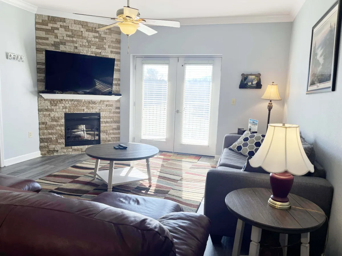 This image features a welcoming living room anchored by a stone fireplace and a prominent flat-screen TV. Large double doors open to the outside, inviting natural light and mountain views into the cozy space. The room is furnished with a comfortable sofa and chairs, creating an inviting area for relaxation and group gatherings.