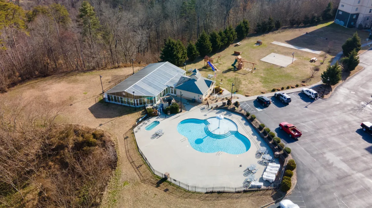 The scene showcases an outdoor recreation area featuring a swimming pool, a playground with colorful equipment, and a building with a glass enclosure in the background. Families and children are seen playing and relaxing, illustrating a lively communal space.
