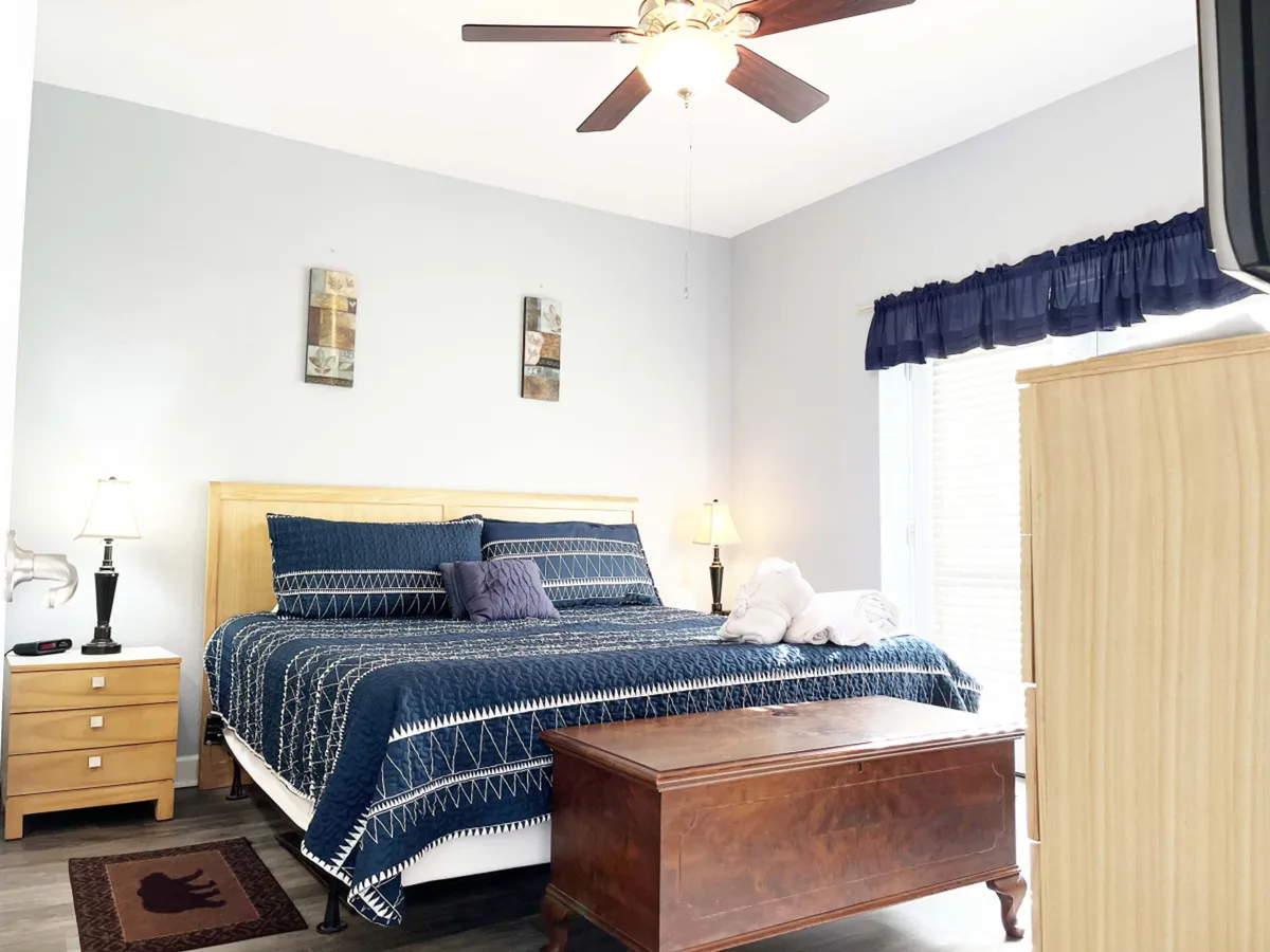 The image features a beautifully made bed with plush linens, a cozy nightstand with a reading lamp, a chest of drawers, and tasteful mountain-inspired decor. Sunlight filters softly through the window, creating a warm, inviting atmosphere ideal for restful mornings and peaceful evenings.